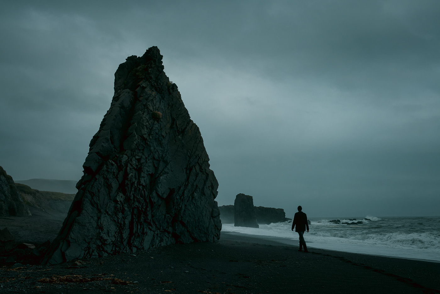 Man walking on black sand beach in Iceland with dramatic clouds over stormy sea.