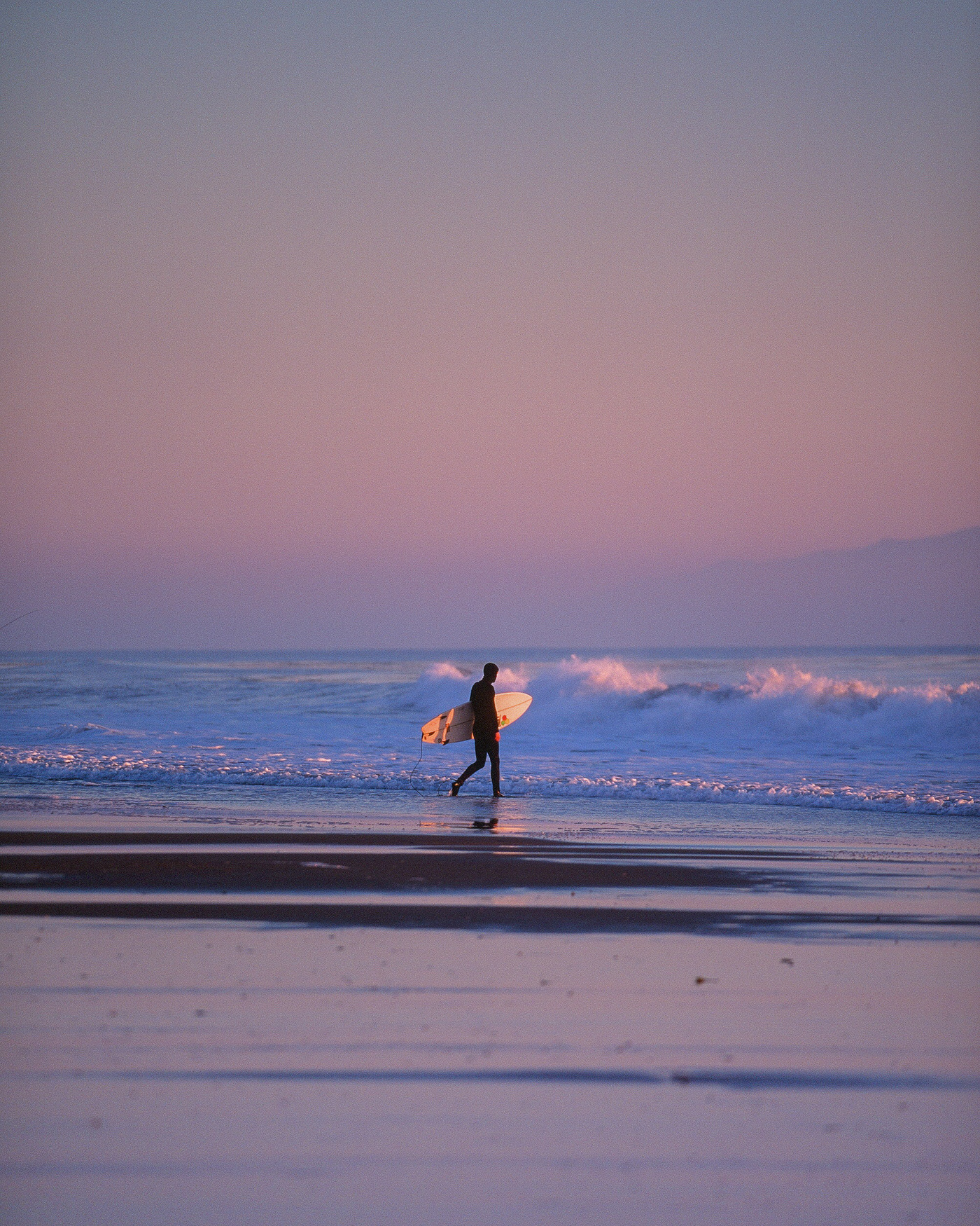 A surfer walks into the ocean at sunset