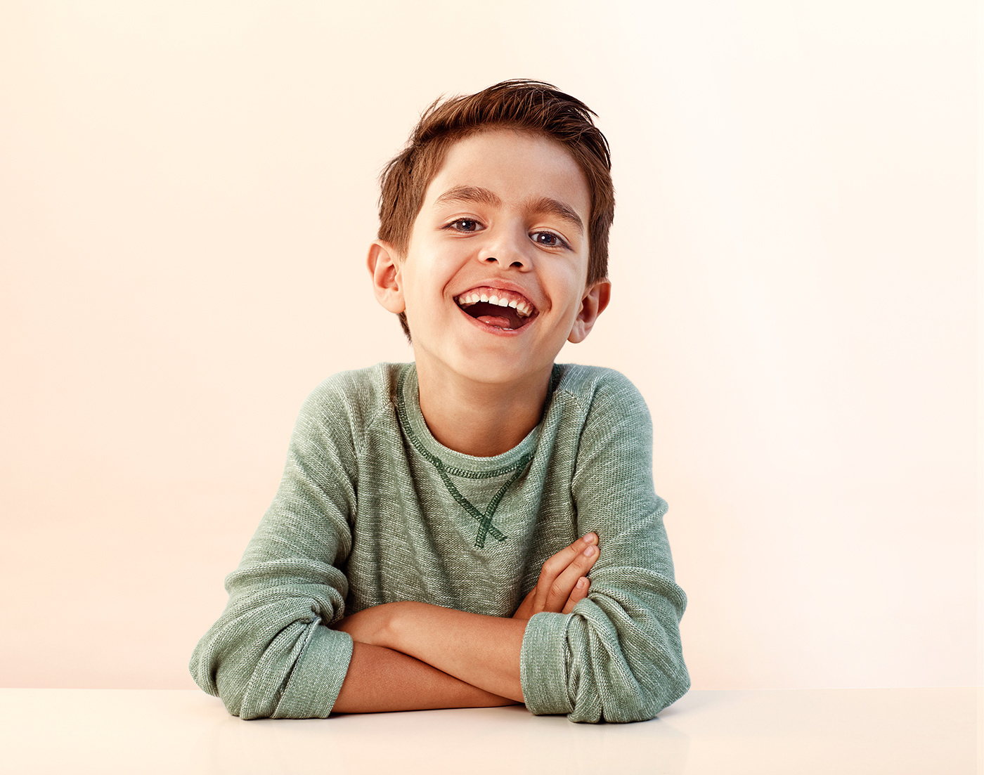 Laughing boy with brown eyes and hair in green shirt with arms crossed.