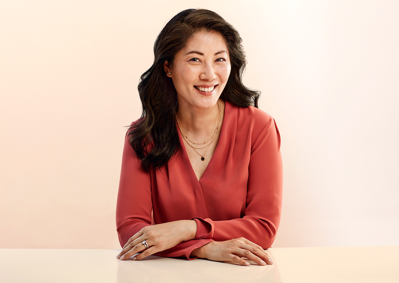 Asian American woman with wavy hair smiling wearing light red shirt, hands folded on table