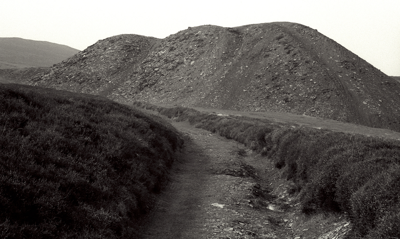 Monochrome image of a grass edged dirt track running beside a slag heap
