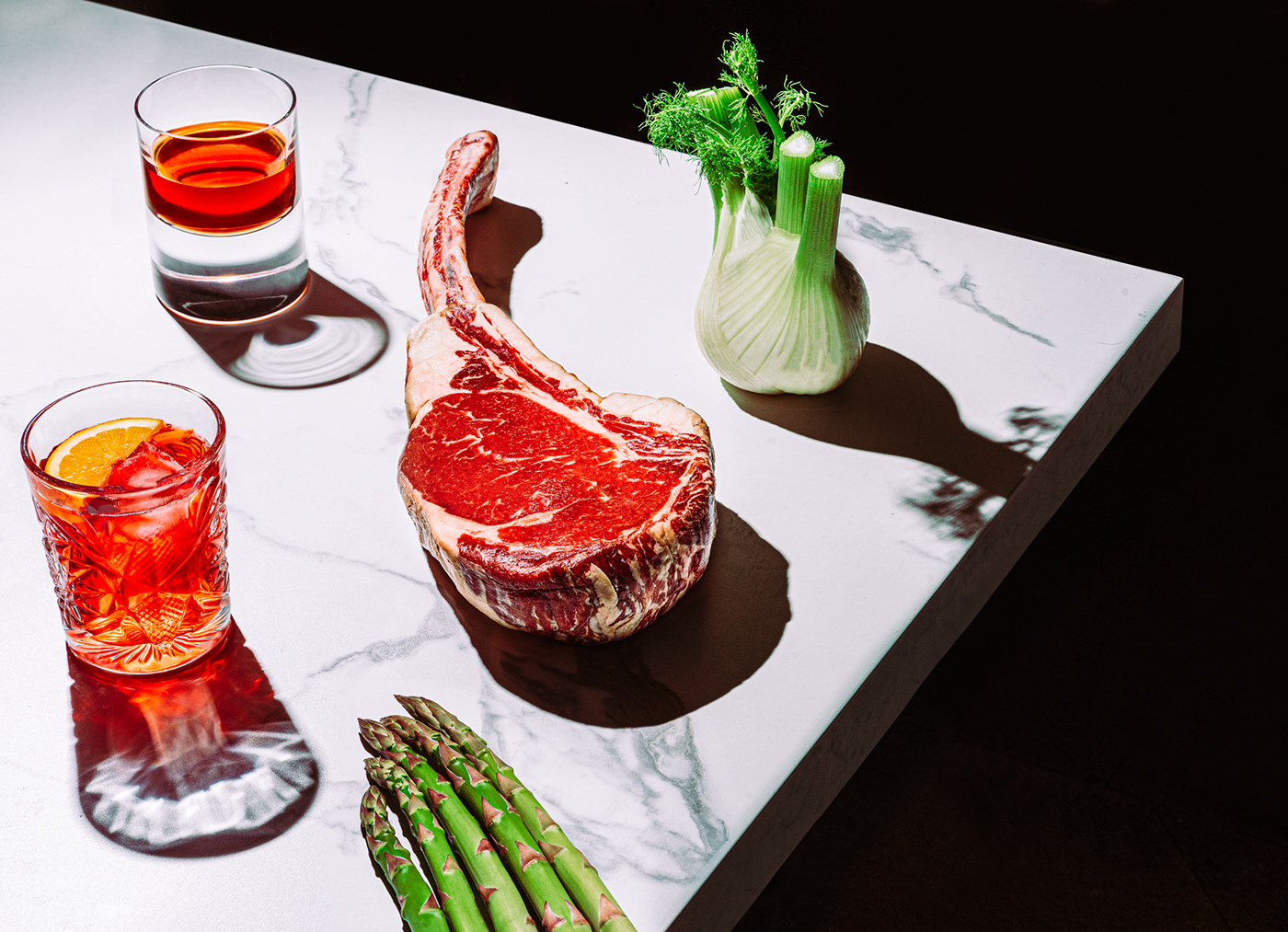 a still life photo of a tomahawk stead with two drinks and vegetables