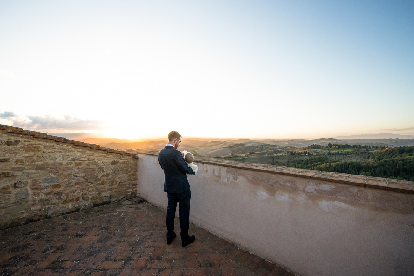 landscape image of the groom on the castle tarrace in front of the sunset. He is holding his baby,