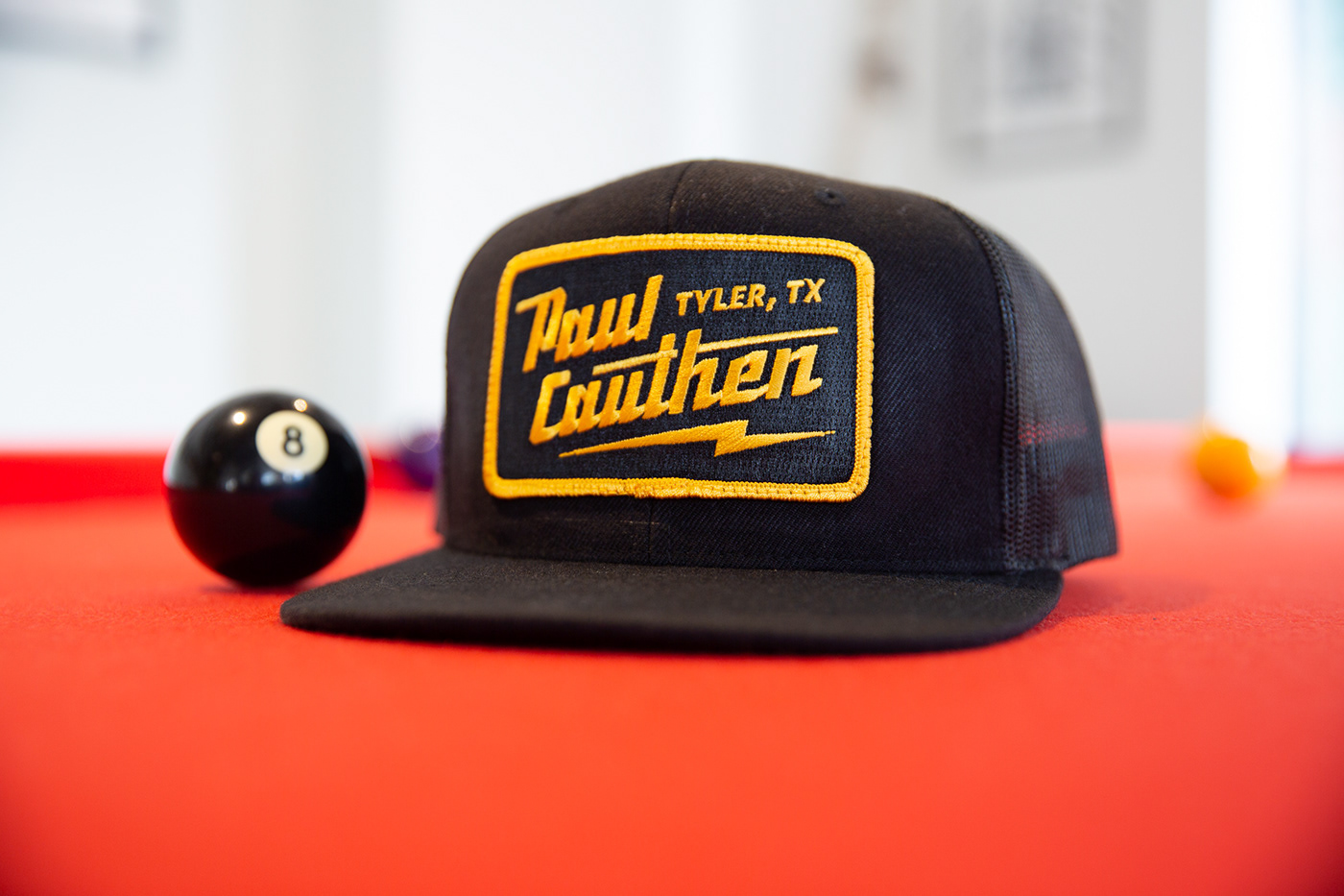 Black trucker hat with an embroidered patch on a red pool table.