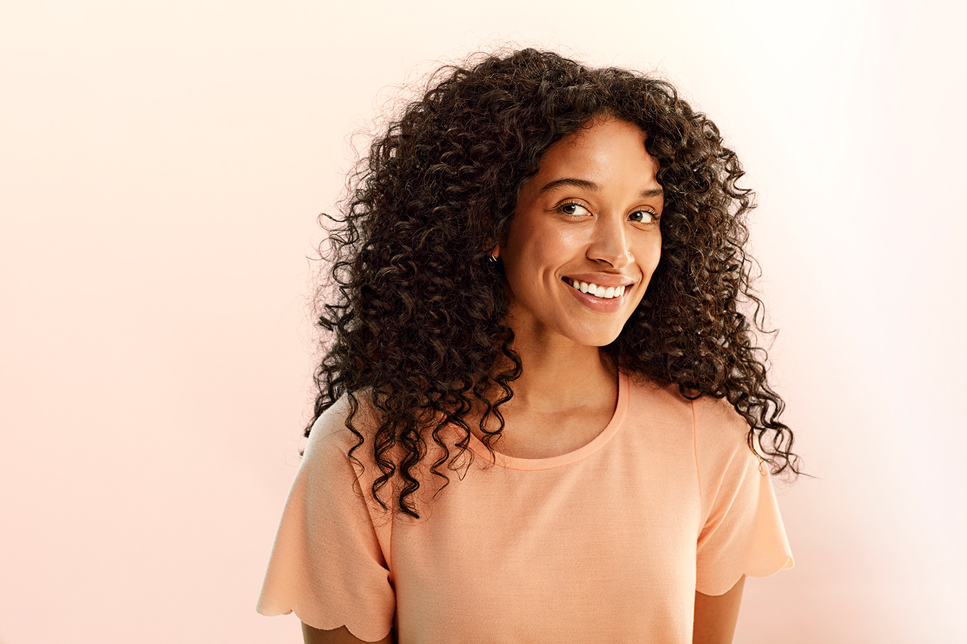 Smiling black woman in peach colored short sleeve shirt. Light cream background.
