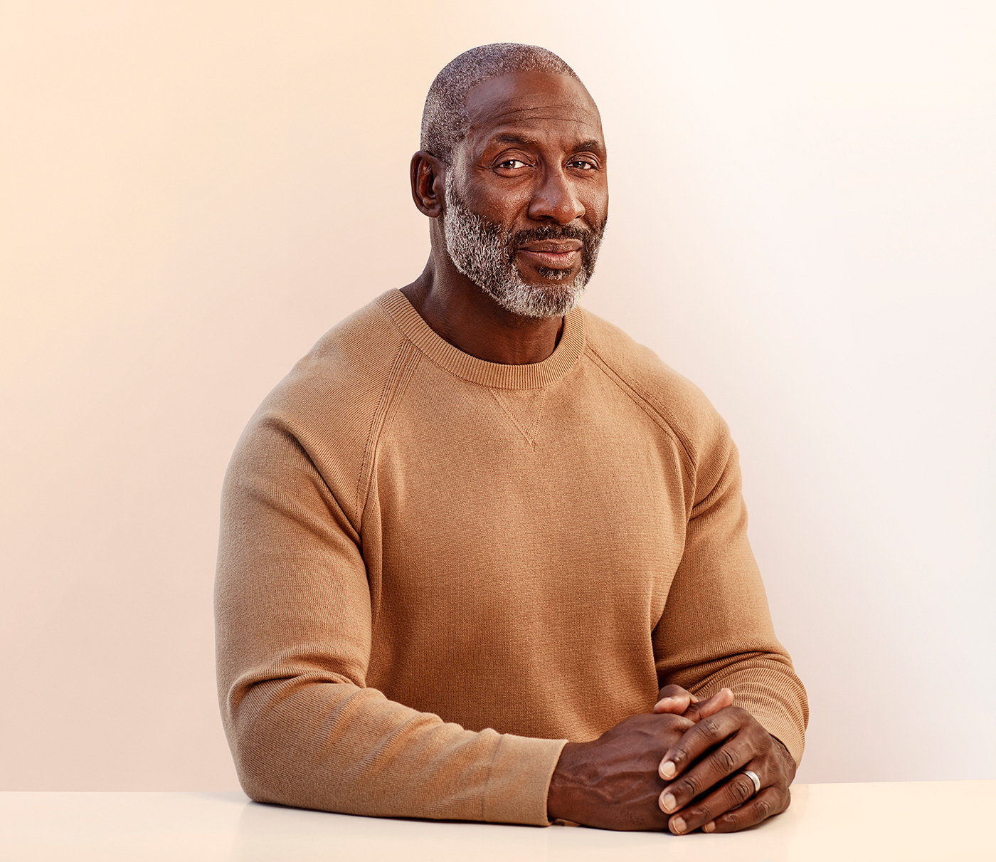 Black man in neutral tan sweater with hands folded gazing straight at camera with neutral expression
