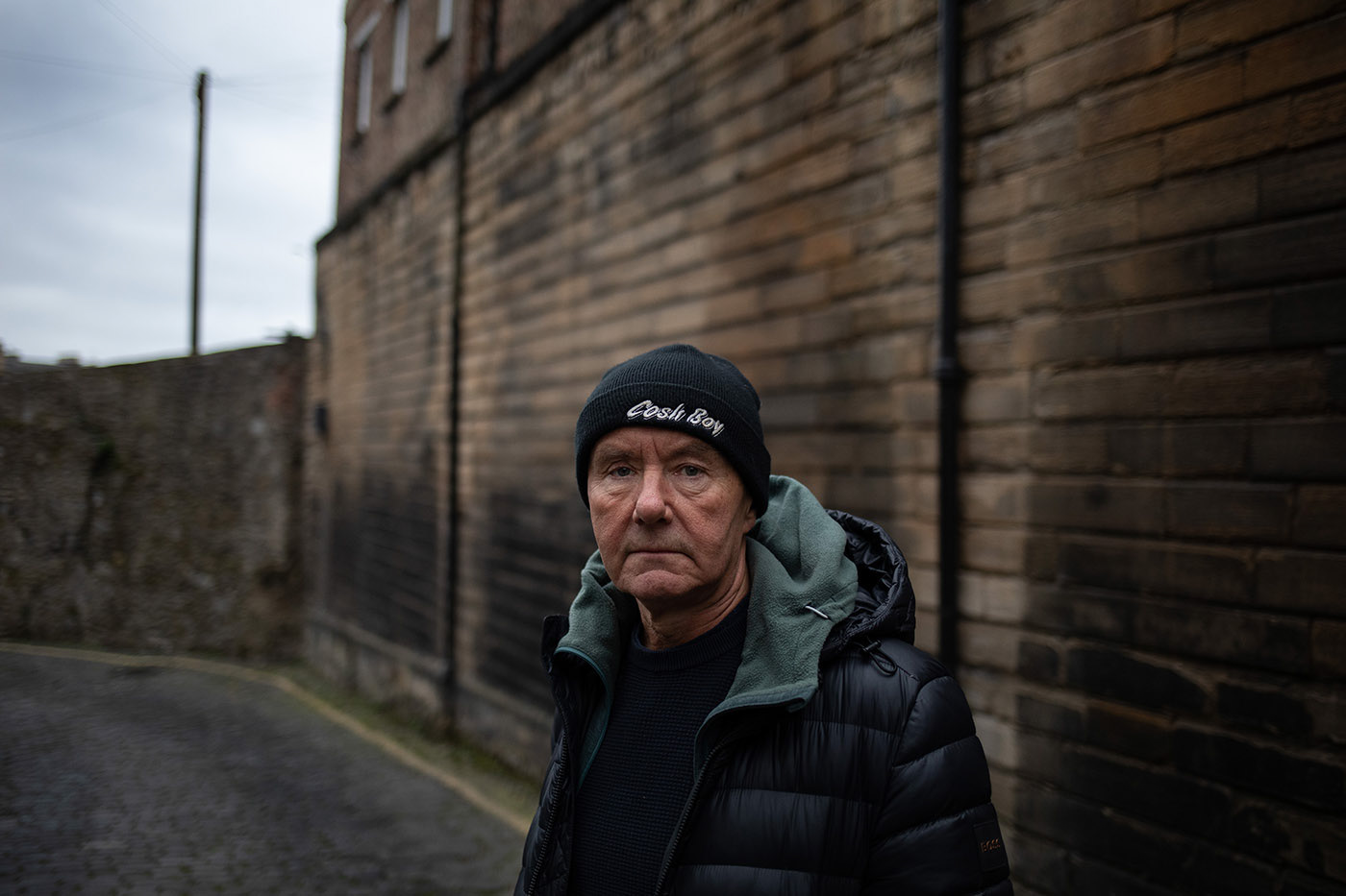 Portrait of crime author Irvine Welsh standing beside tall wall in a moody lane.