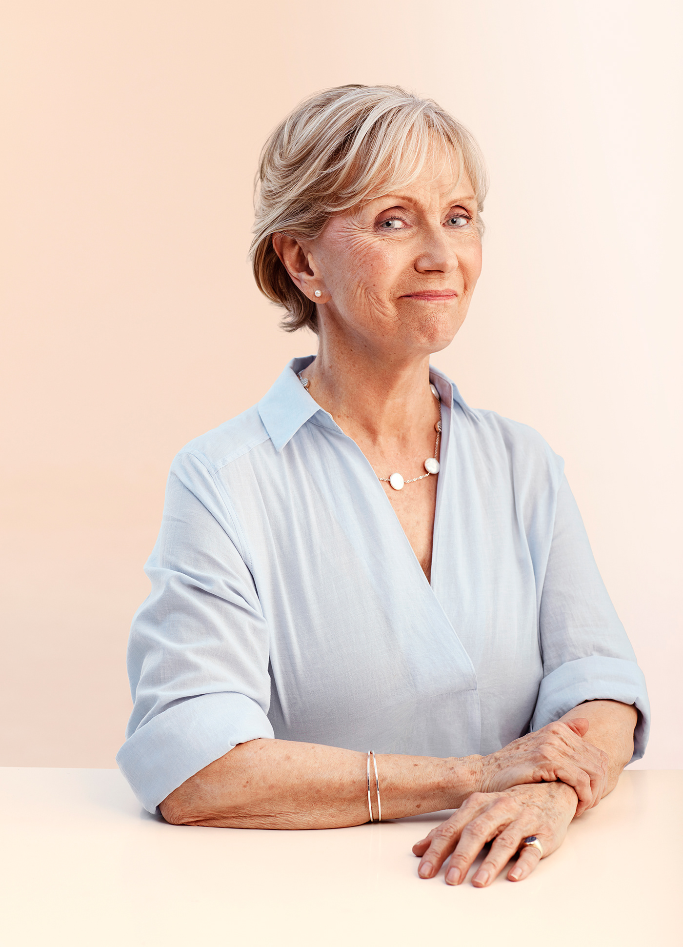 Smiling older woman with short blond hair, wearing light blue collared shirt. Arms folded on table. 