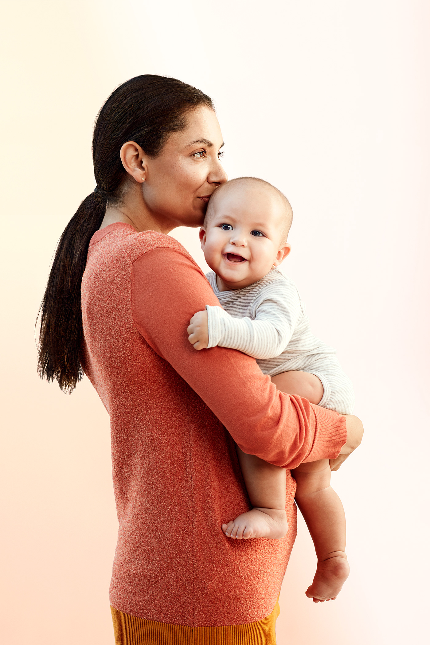 Mother with long dark hair in low-ponytail holding smiling baby in arms, kissing baby's head. 