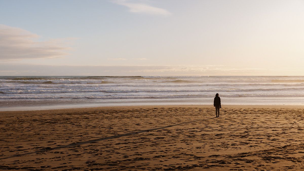 A silhouette of a woman standing on a golden sand beach looking towards the sun at golden hour.