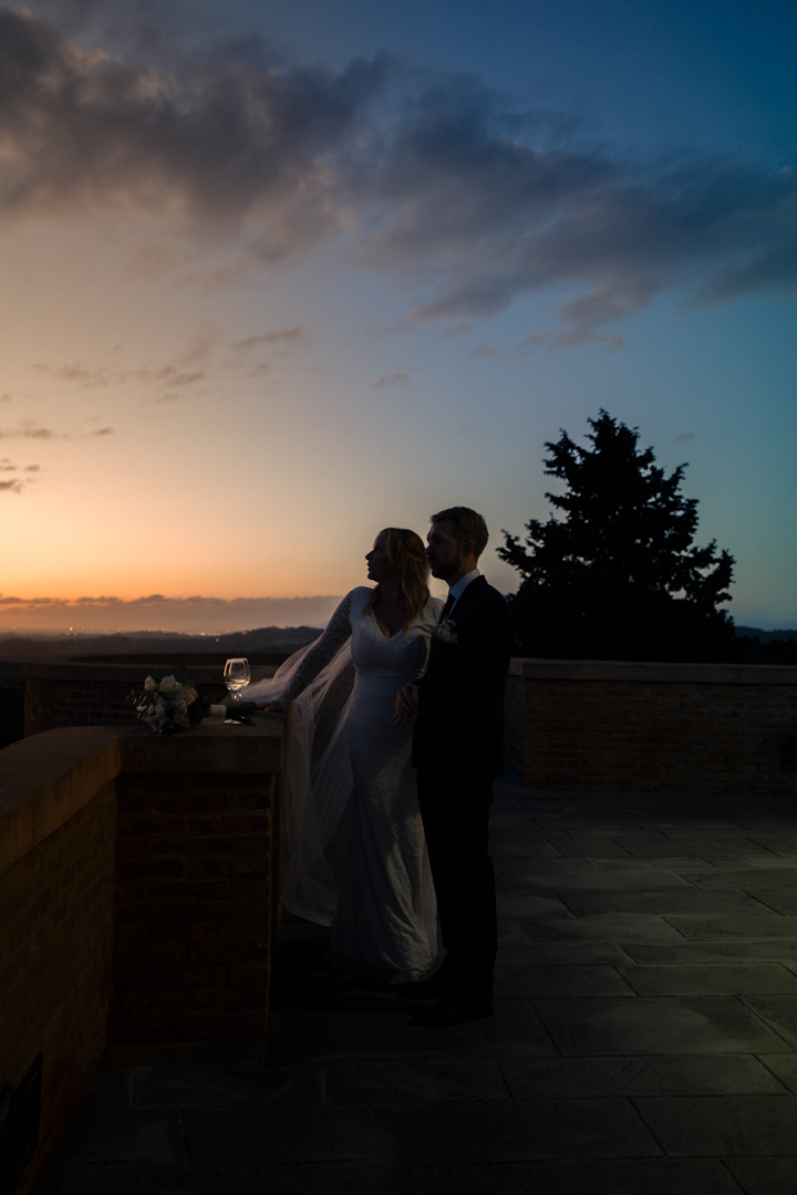 Bride and groom on a terrace in front of the sunset drinking wine after their wedding. 
