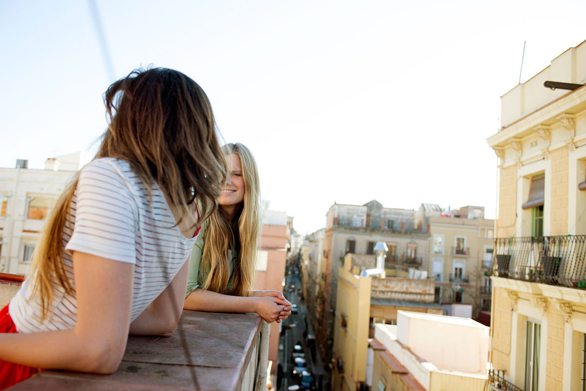 Two women are chatting on a rooftop with view on the city, roofs and streets.