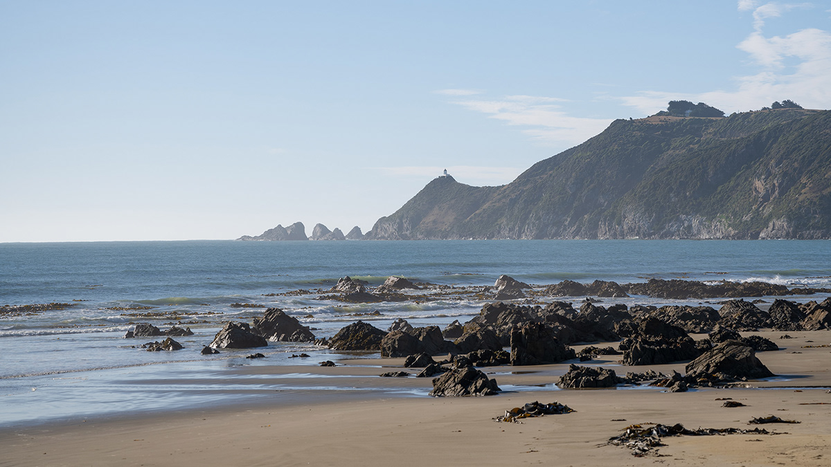 An ocean beach with soft golden sand and the Nugget Point Lighthouse in the distance.