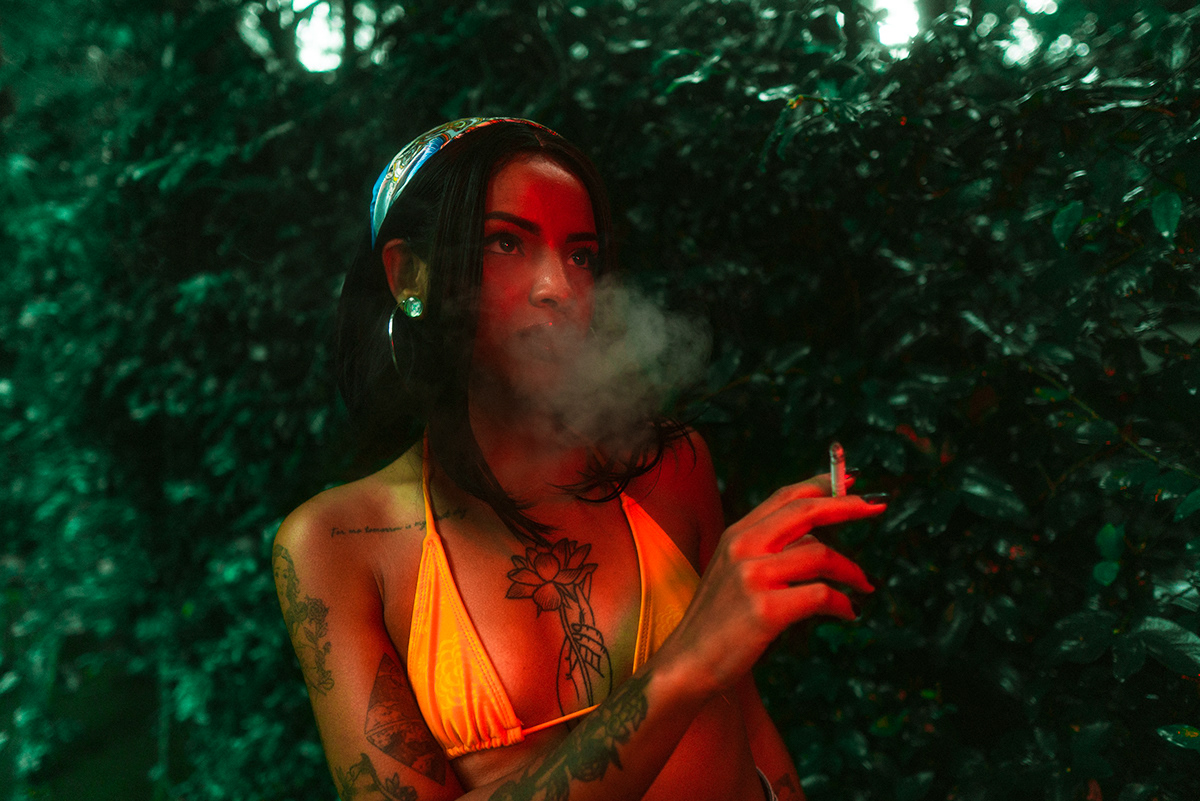Garota iluminada por luz vermelha no meio de um ambiente de natureza, segurando um cigarro.