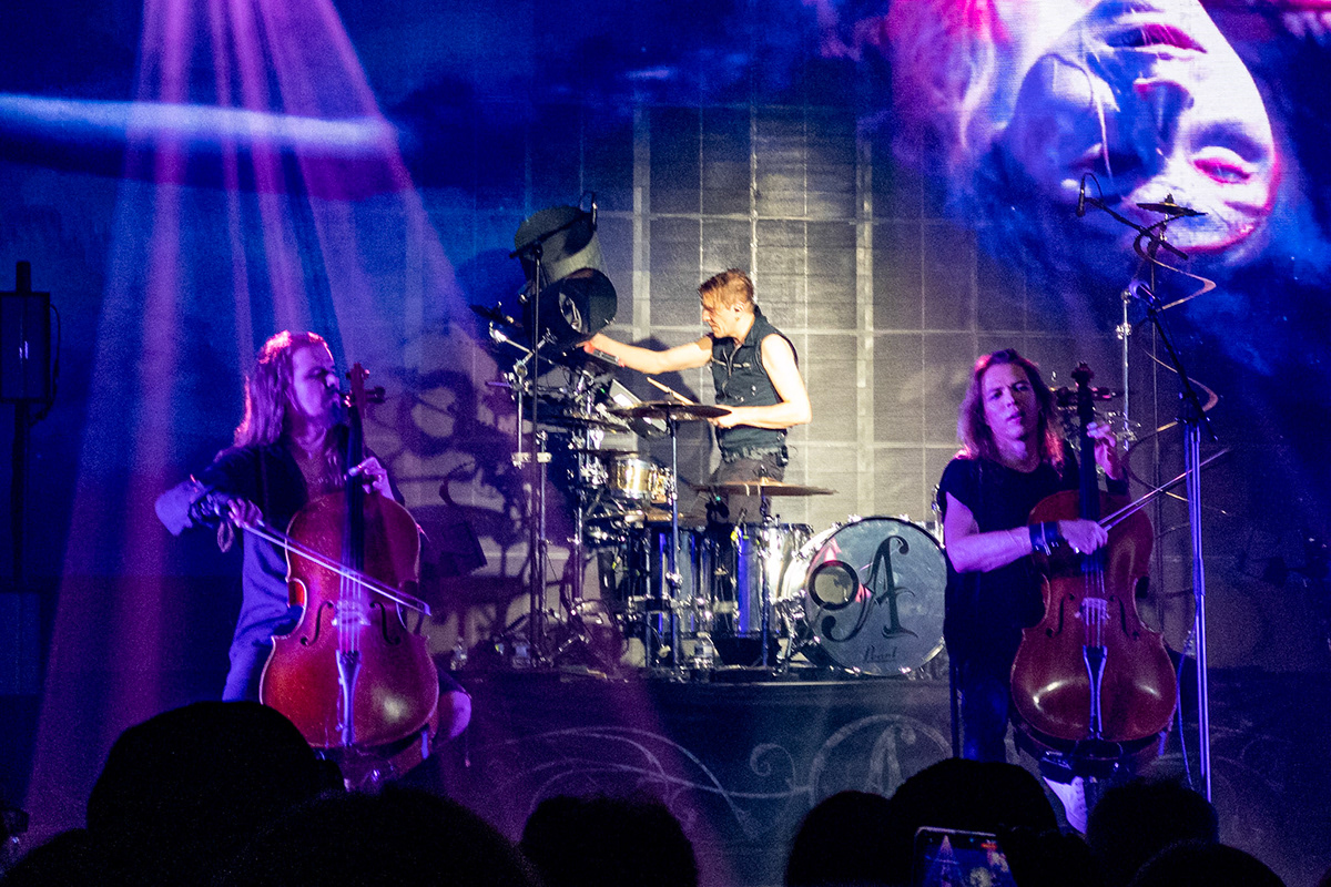 Two men playing rock/metal cello on stage with a man playing drums in the background