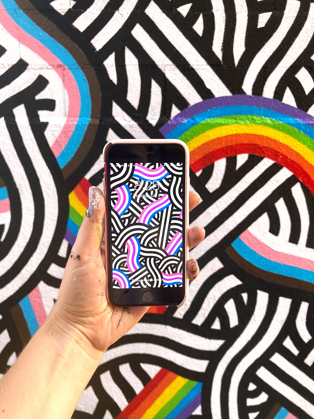 A hand holding a phone with a pattern showing the bisexual flag colors in front of a pride mural.