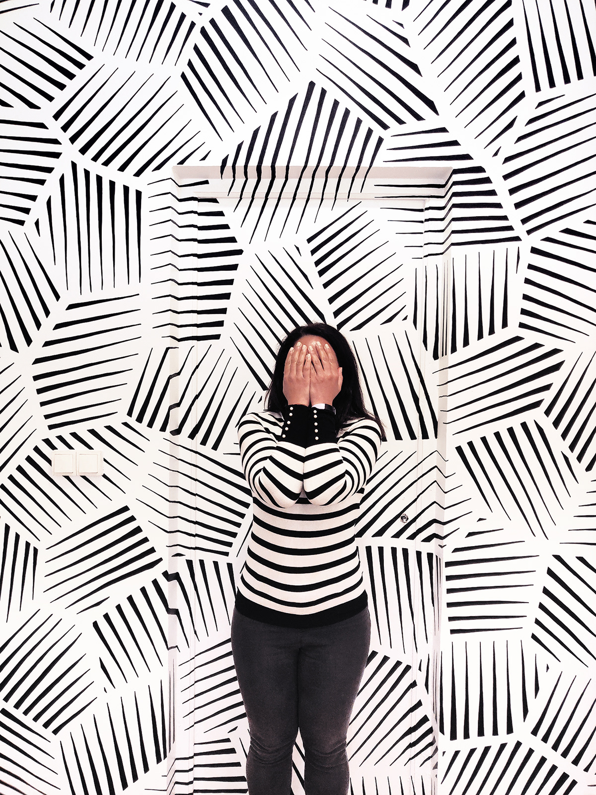 A woman camouflaging in front of an abstract, geometric black and white mural.